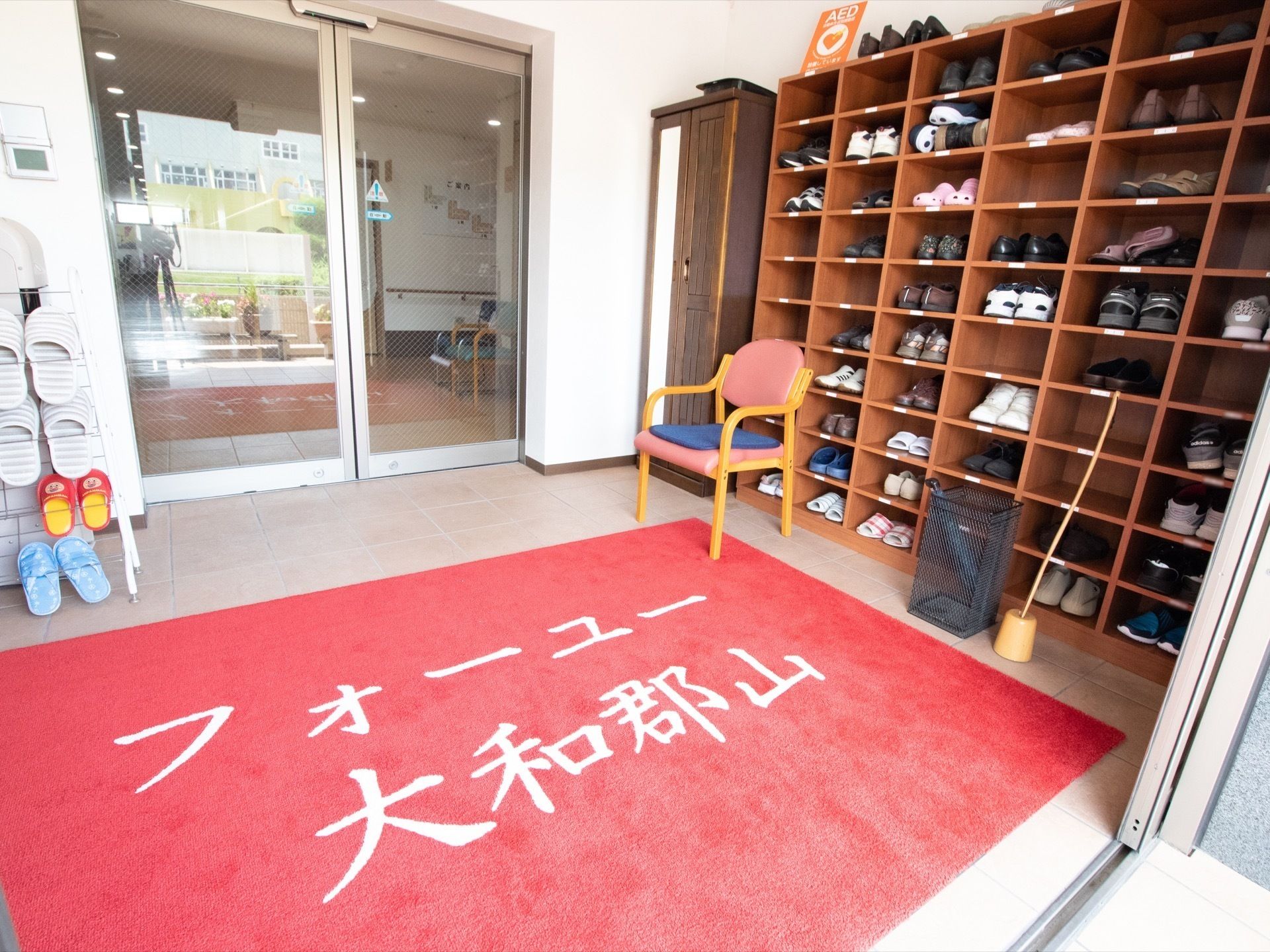 白色の文字で施設名称がかかれた大きな赤色のマットが敷かれている、外玄関を入った所にある土間
