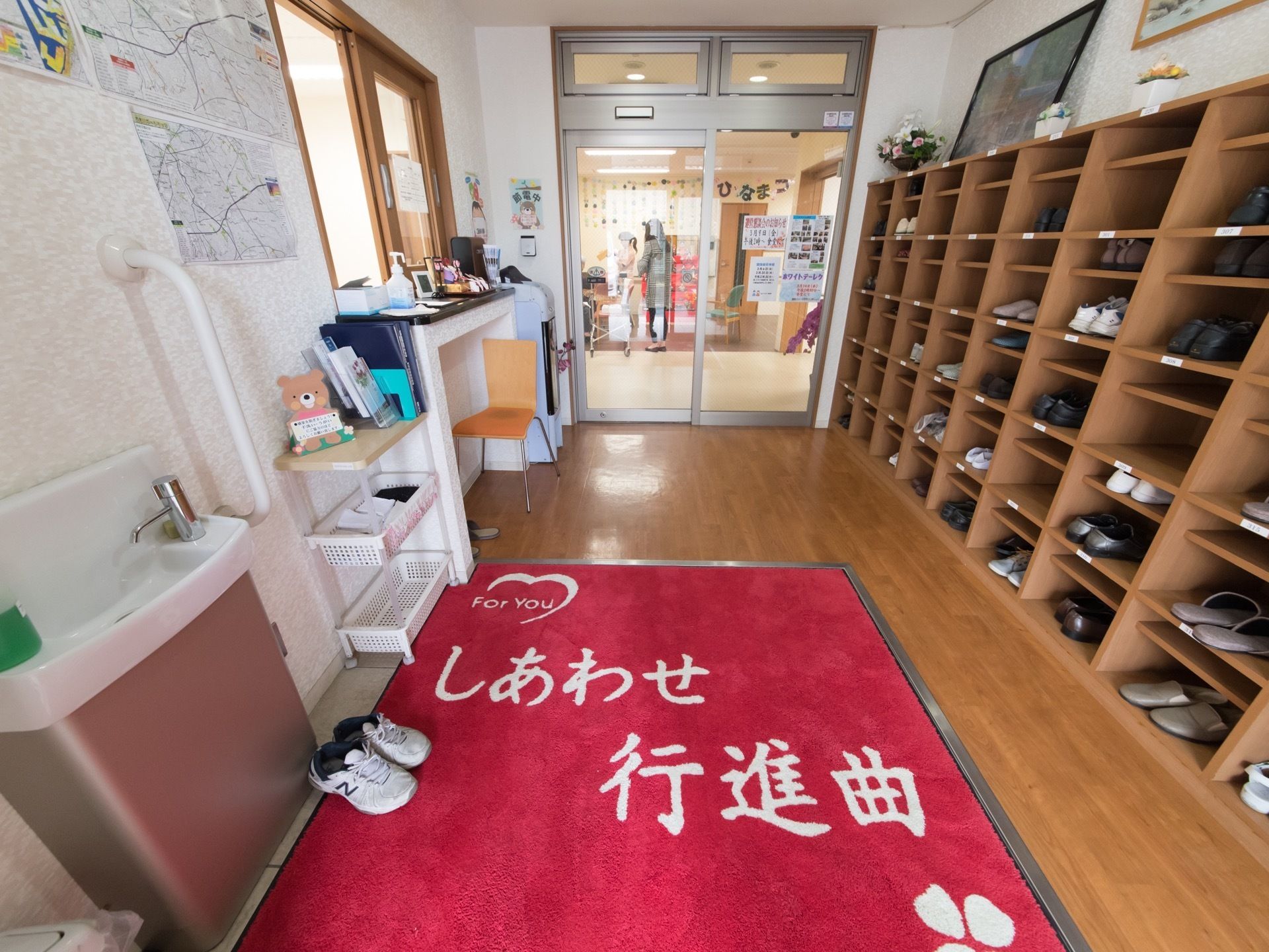 洗面台や靴箱、受付の小窓のある風除室である。足元には赤字の白い文字の書かれた玄関マットが敷かれている。