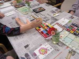 新聞紙を広げたテーブルの上で絵手紙を書いている入居者たち。様々な形のパレットがある。