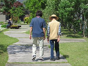 紺の服を着ているスタッフが帽子をかぶった高齢男性に付き添って、庭を歩いている。