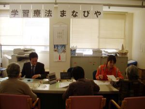 男女の先生のような人が机についていて、対面式で3名の高齢女性たちが座り、プリントをしている。