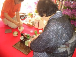 グレーの着物を着て抹茶を立てている高齢女性。本格的な抹茶セットがあり、オレンジのポロシャツを着た女性スタッフがいる。
