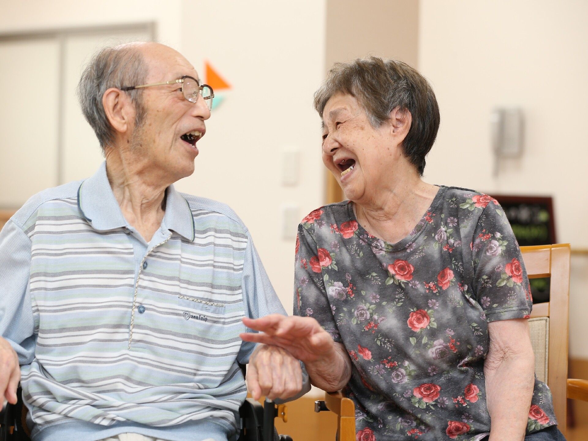 眼鏡をかけた高齢男性と花柄の服を着た高齢女性が隣同士に座り、楽しそうに話をしている。