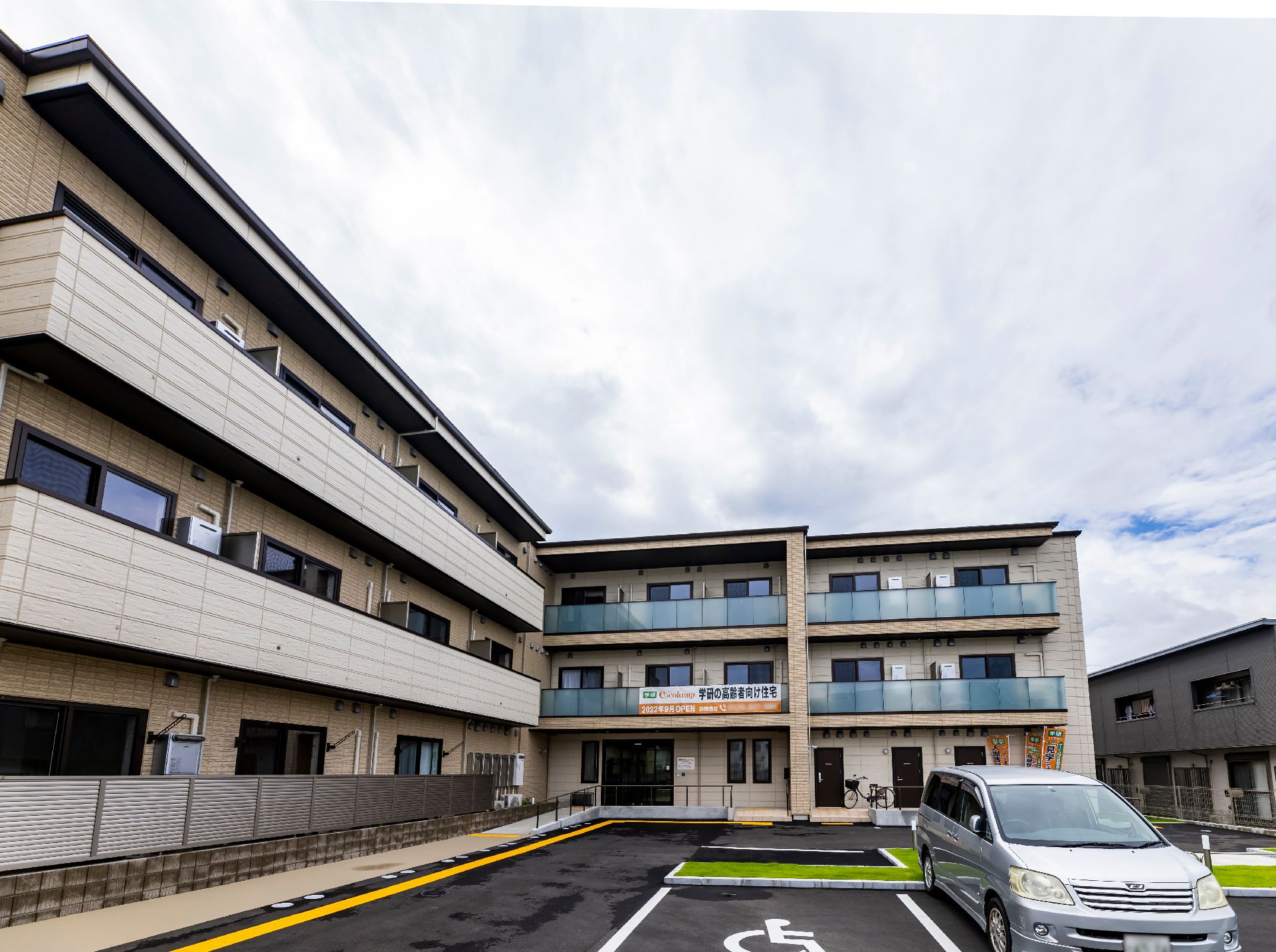 車が止まっている駐車場に隣接したベージュの外観で３階建ての建物