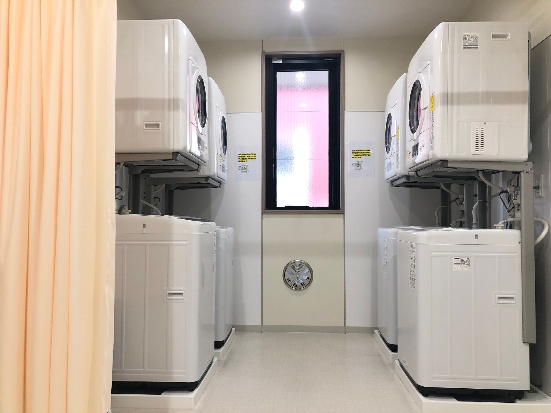 縦型の洗濯機の上に乾燥機が置かれたセットが４組置かれている。ピンクのカーテンで仕切られている。