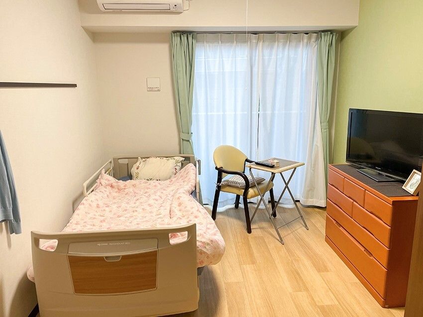 寝具付きのベッドやタンス、テレビなどが置かれたフローリングの部屋には大きな窓が付いている。