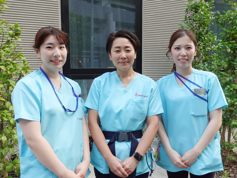 自然が広がる場所で車いすに乗りリラックスする女性とスタッフ。カメラに笑顔を向けている様子