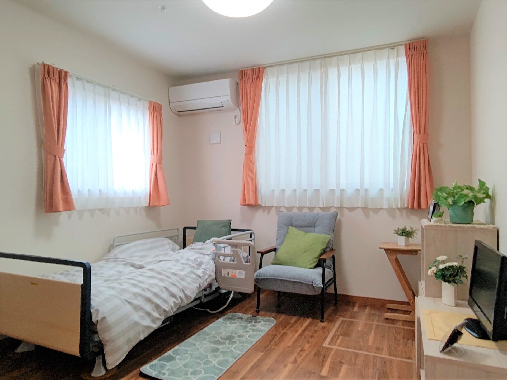 草花が飾られた部屋には介護ベッドやいす、テレビなどがあり、壁にはエアコンが設置されている。
