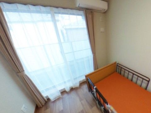 オレンジの介護ベッドが置かれたフローリングの部屋にはカーテン付きの大きな窓やエアコンがある。