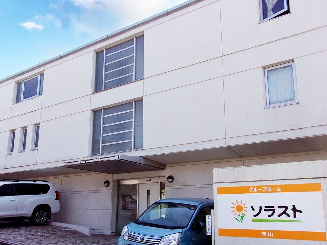 緑の多い屋外で笑顔で過ごしている車いすの高齢女性と付き添いのピンクのエプロンを付けた若い女性スタッフ。