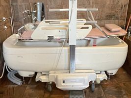 ストレッチャータイプのリフトがある大きな浴槽の機会。白とピンクの寝台浴が設置されている。