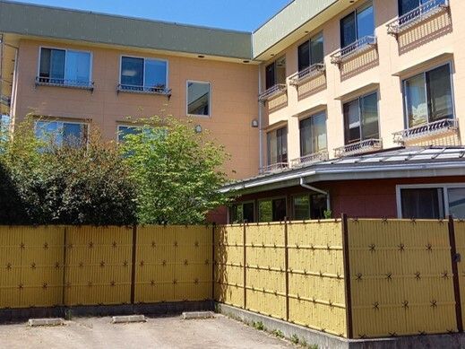 駐車場が設置されている敷地内には3階建ての建物がありブラウン系の外壁。建物の周りには草木が植えられている。
