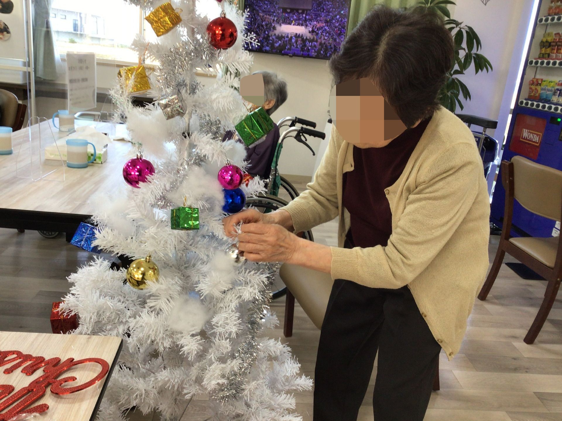 高齢女性が共有スペースにある白いクリスマスツリーの飾りつけをしている様子。