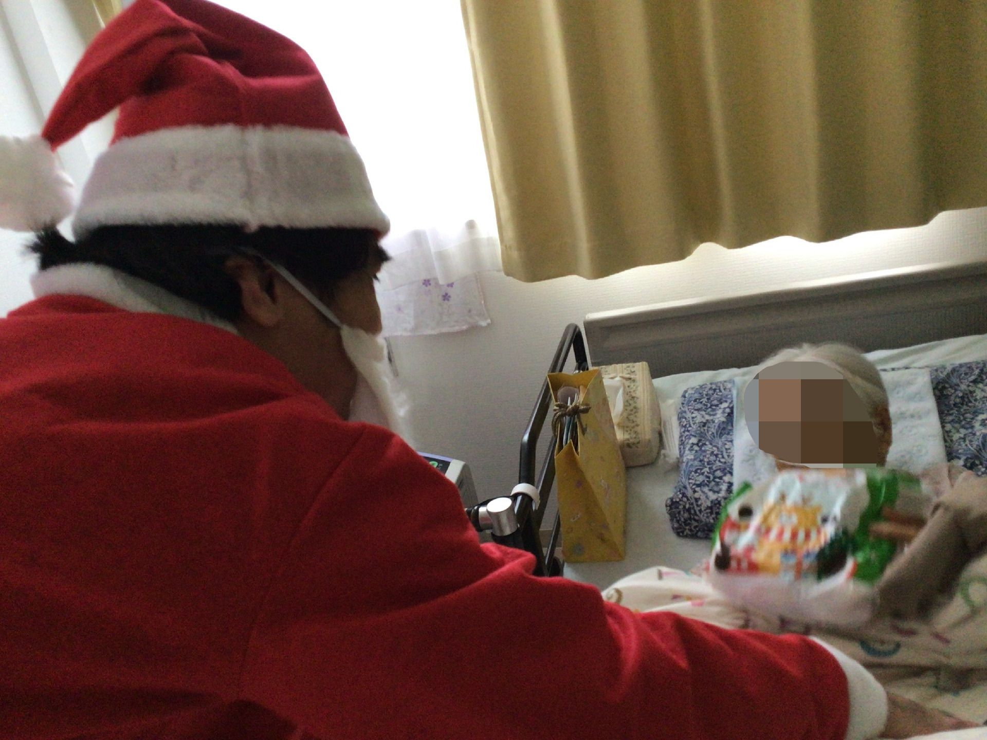 サンタ帽をかぶって髭を付け、赤いサンタクロースの服を着たスタッフがベッドで寝ている高齢女性にプレゼントを渡している。