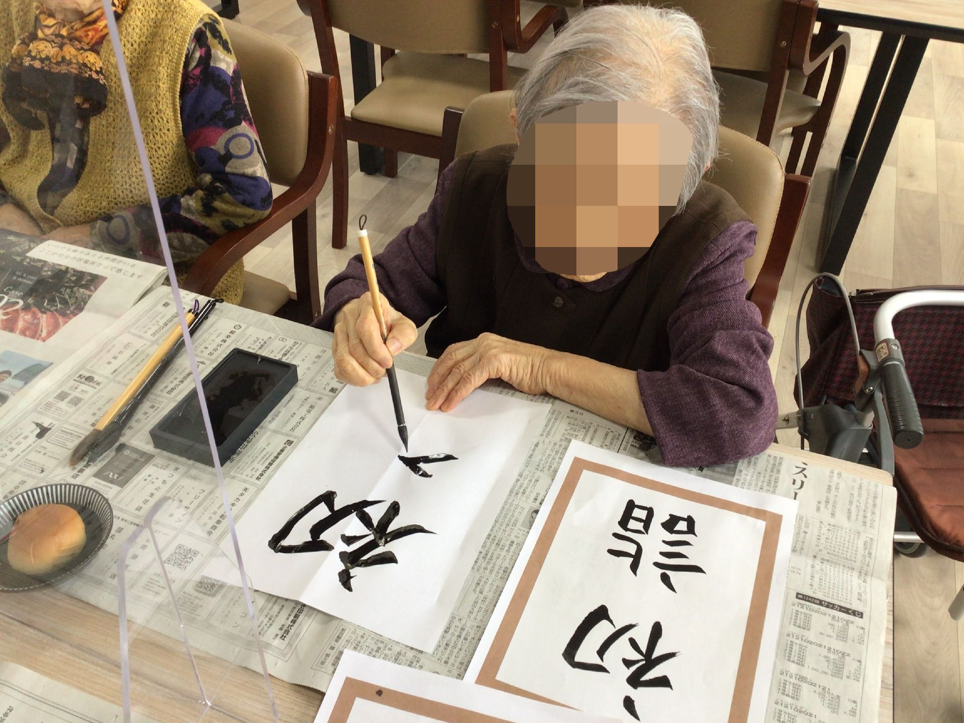新聞が敷かれているテーブルの上には半紙が置かれ、高齢女性が見本を見ながら習字をしている。