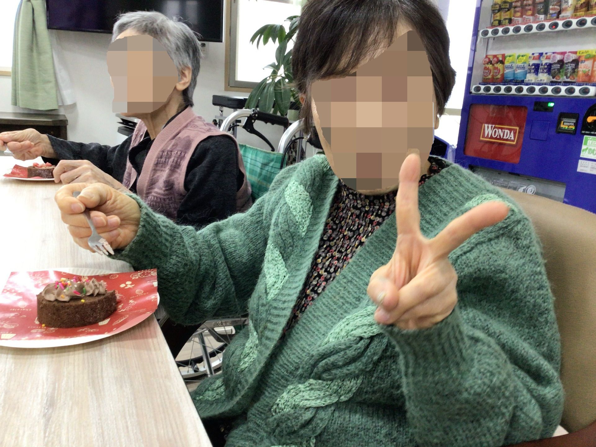自動販売機が設置されている食堂のテーブルにはチョコレートケーキがあり、高齢女性が笑顔で食べている。