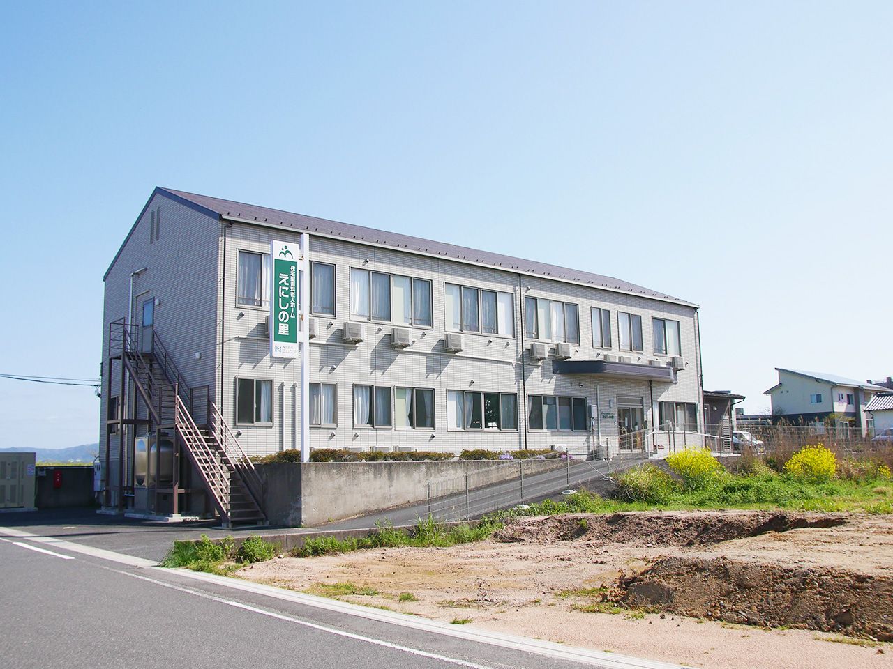 青い空の下に建っている２階建ての建物。周囲には低木が植えられ、建物の前の坂を上る車いすの高齢者と付き添いの人がいる。