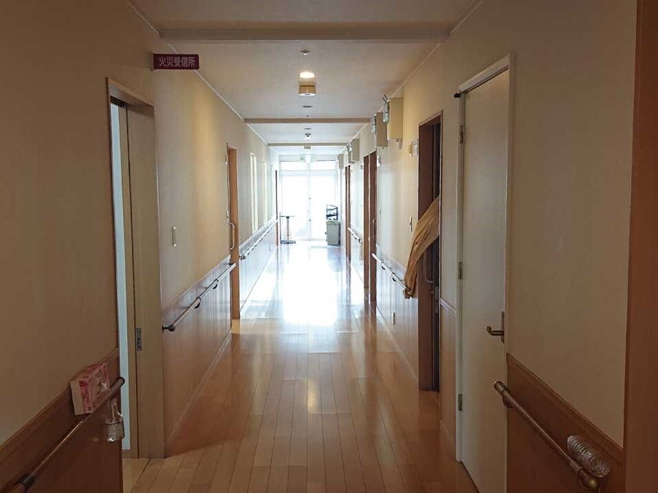 手すりや居室のドアが両サイドに設置されたまっすぐな廊下。突き当りのドアから光が入っている。