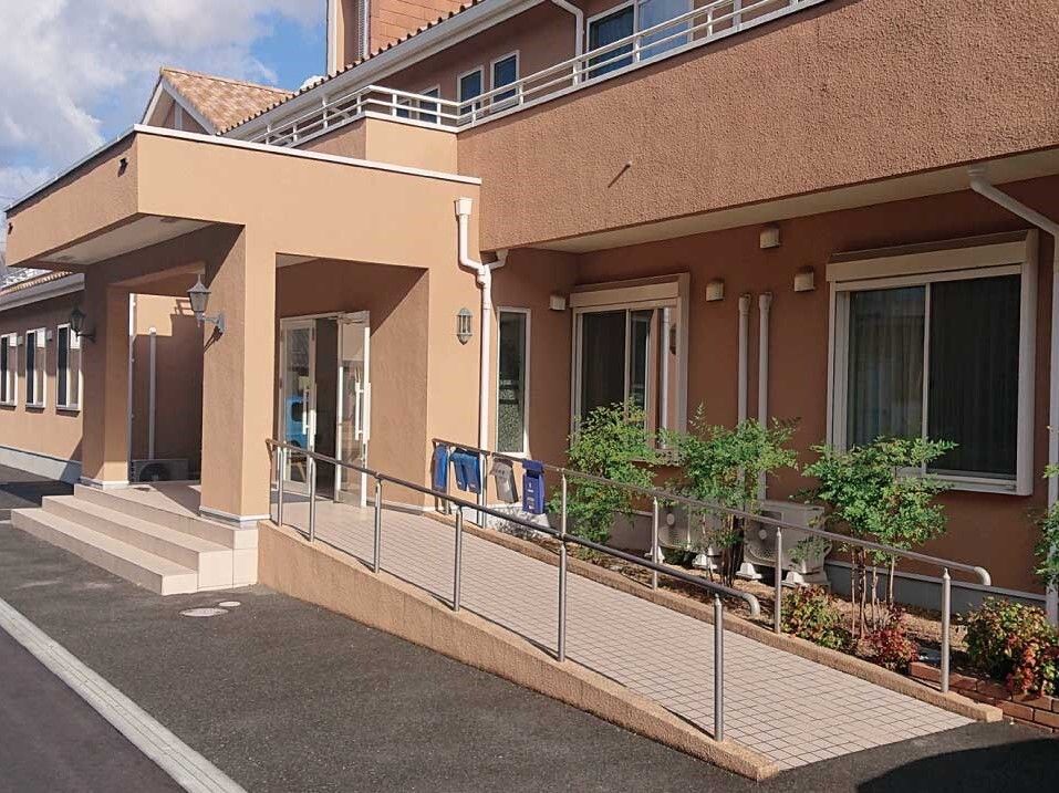 手すり付きのスロープと階段が付いている玄関。ブラウン系の建物は2階建てで草木が植えられている。