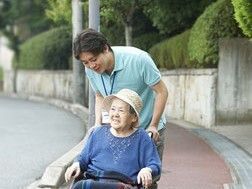 緩やかな坂道を散歩している車いすの高齢女性とポロシャツを着た男性スタッフ。