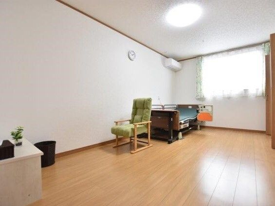 介護ベッドや一人用の椅子がセットされたフローリングの居室。ゴミ箱やエアコン、時計もある。