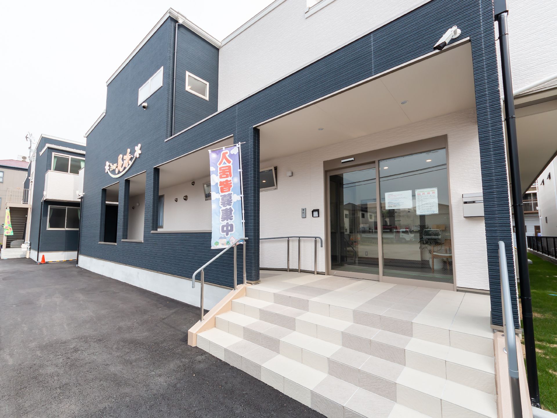 日当たりのいい場所に建っている２階建ての建物。モノトーンでまとめられ、玄関には手すり付きの階段とスロープがある。