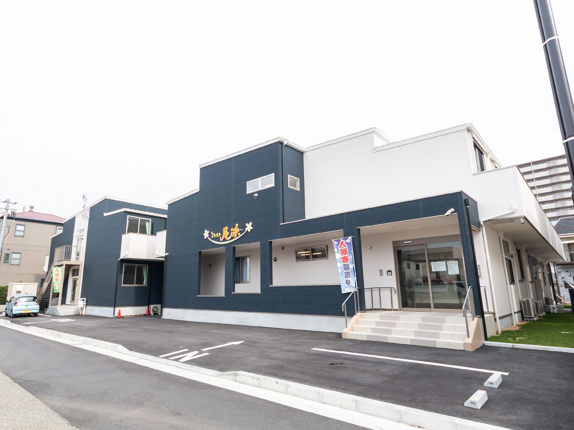 2階建ての建物はダークグレーと白で、玄関には手すり付きの階段とスロープがあり、日当たりのいい場所に建っている。