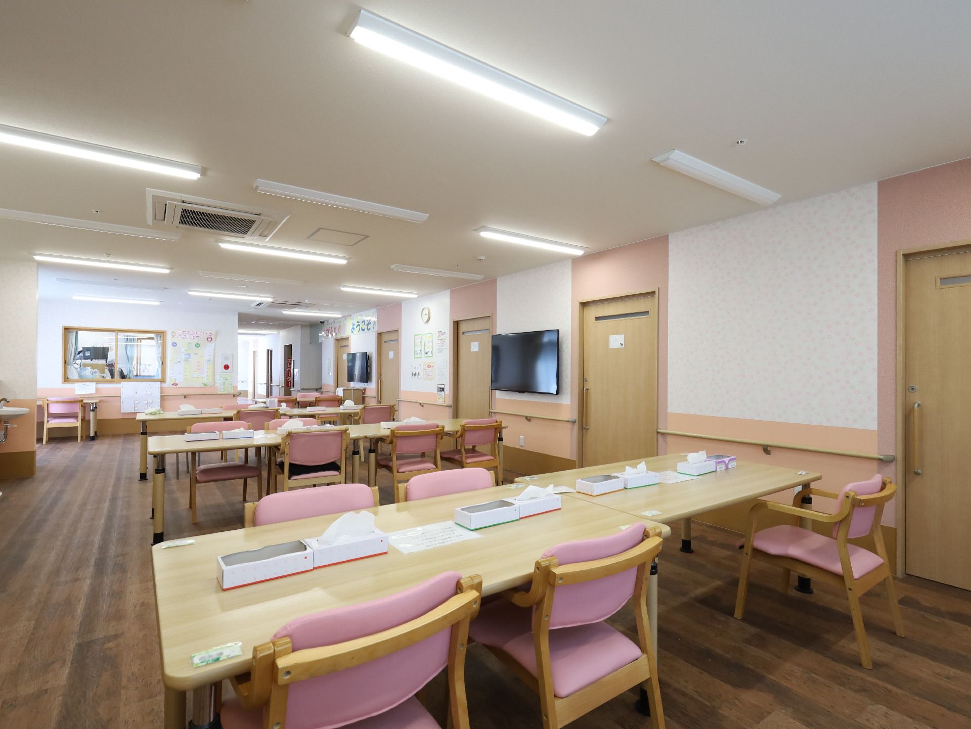 木製の机とピンクのクッションが付いた椅子が配置された白い壁の広間