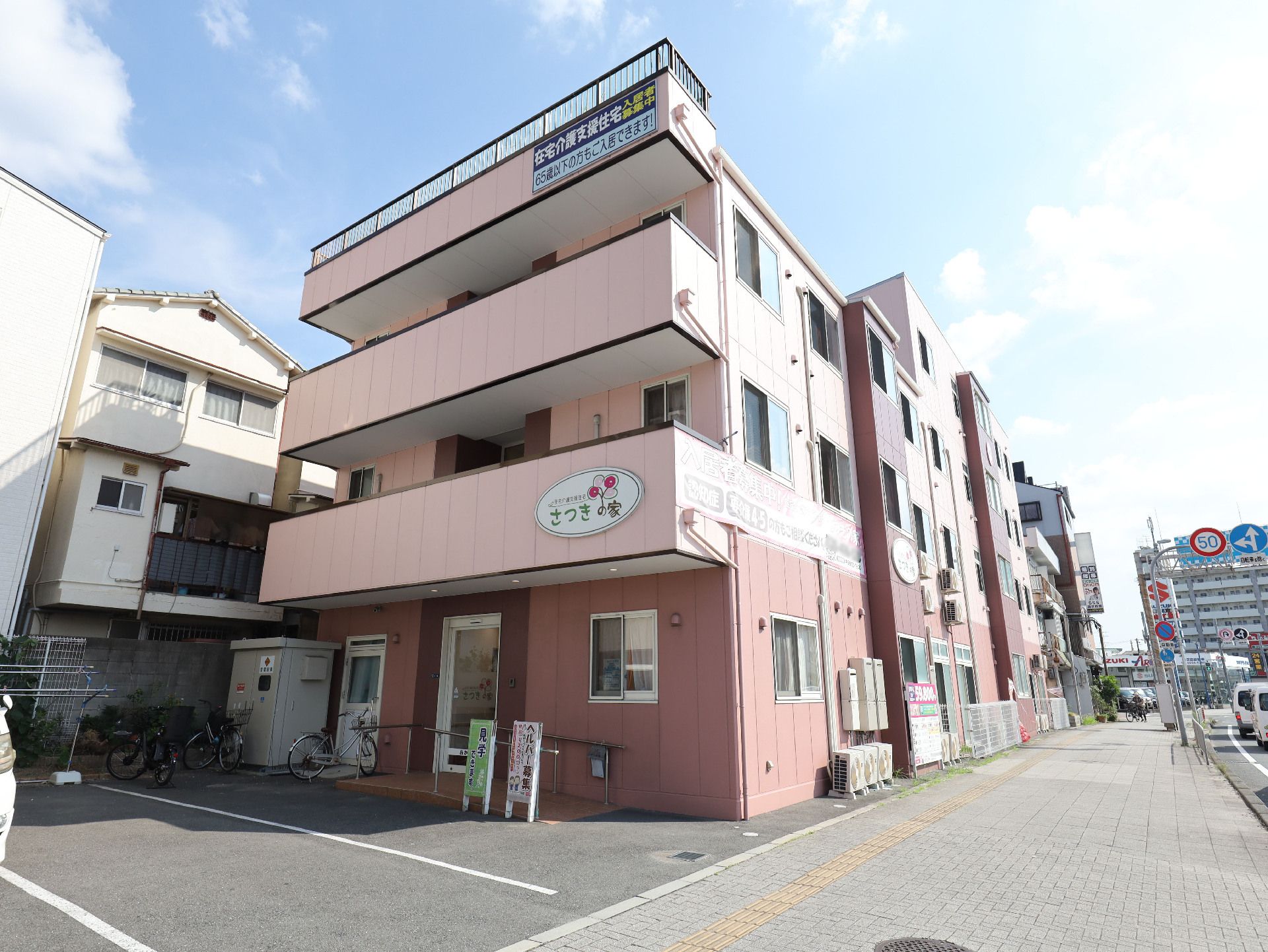 アスファルトの歩道沿いに立地しているピンクの外観でベランダが付いている３階建ての建物