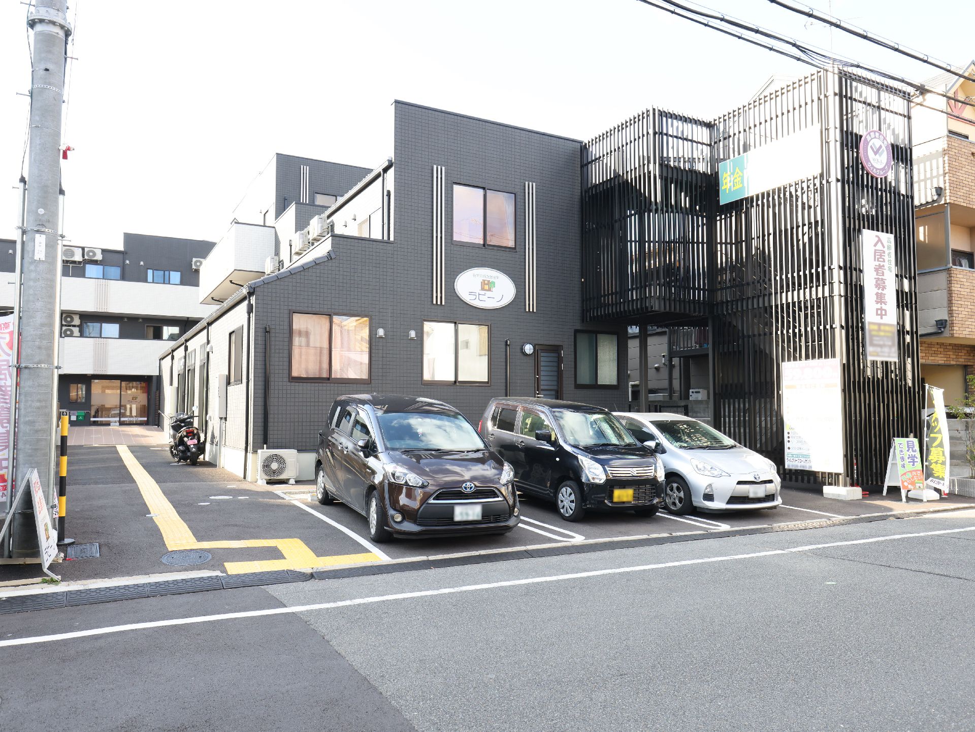 ダークグレーを基調とした３階建ての建物は屋外階段が設置され、敷地内には駐車場が付いている。