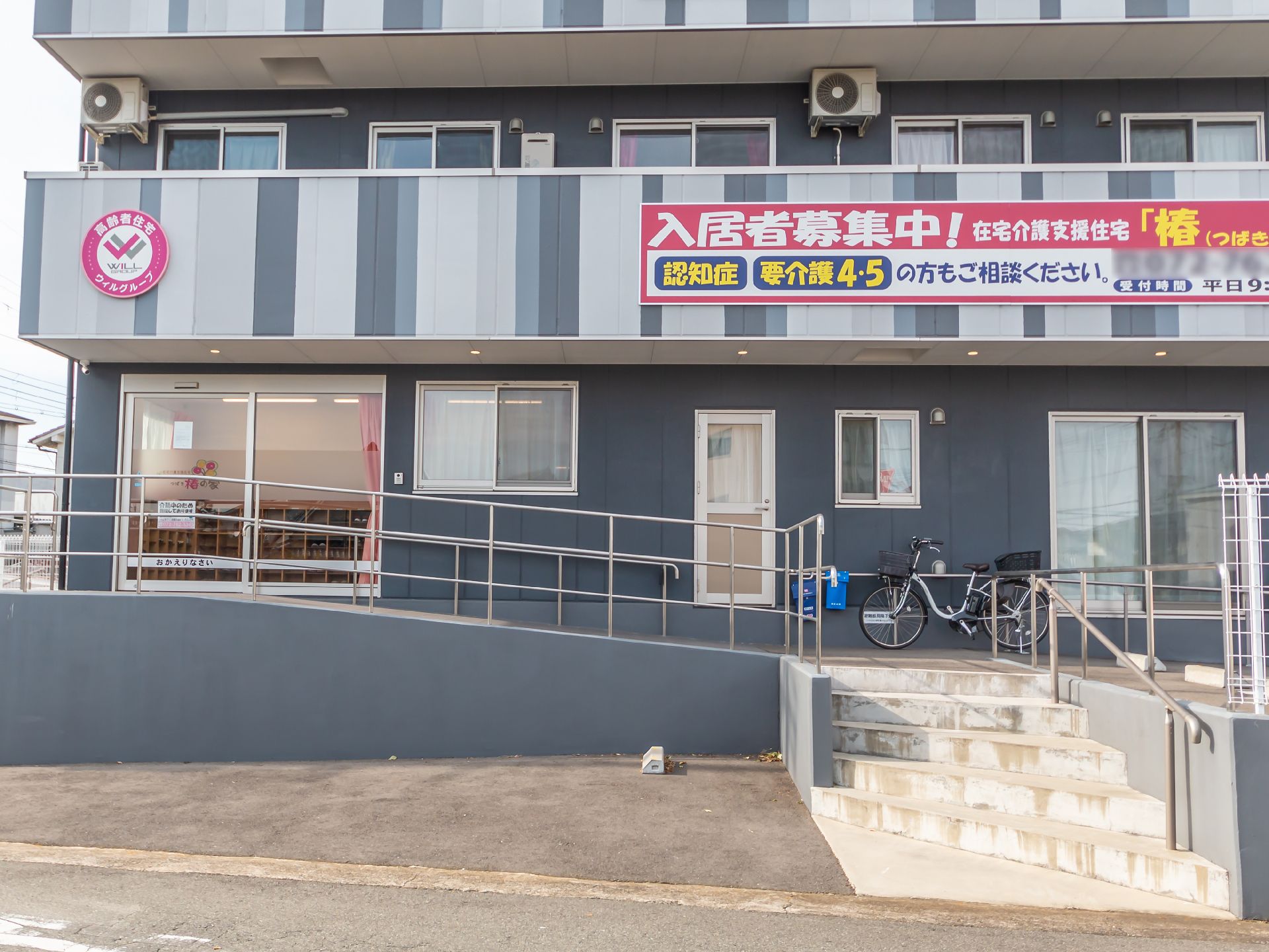 3階建ての建物はグレーと白で周辺には緑があり、敷地内には駐車場がある。草花も植えられている。