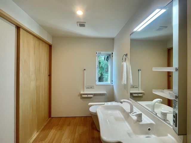 大きな鏡が付いた洗面所はワイドタイプでタオル掛けがそばにあり、奥には手すり付きのトイレがある。
