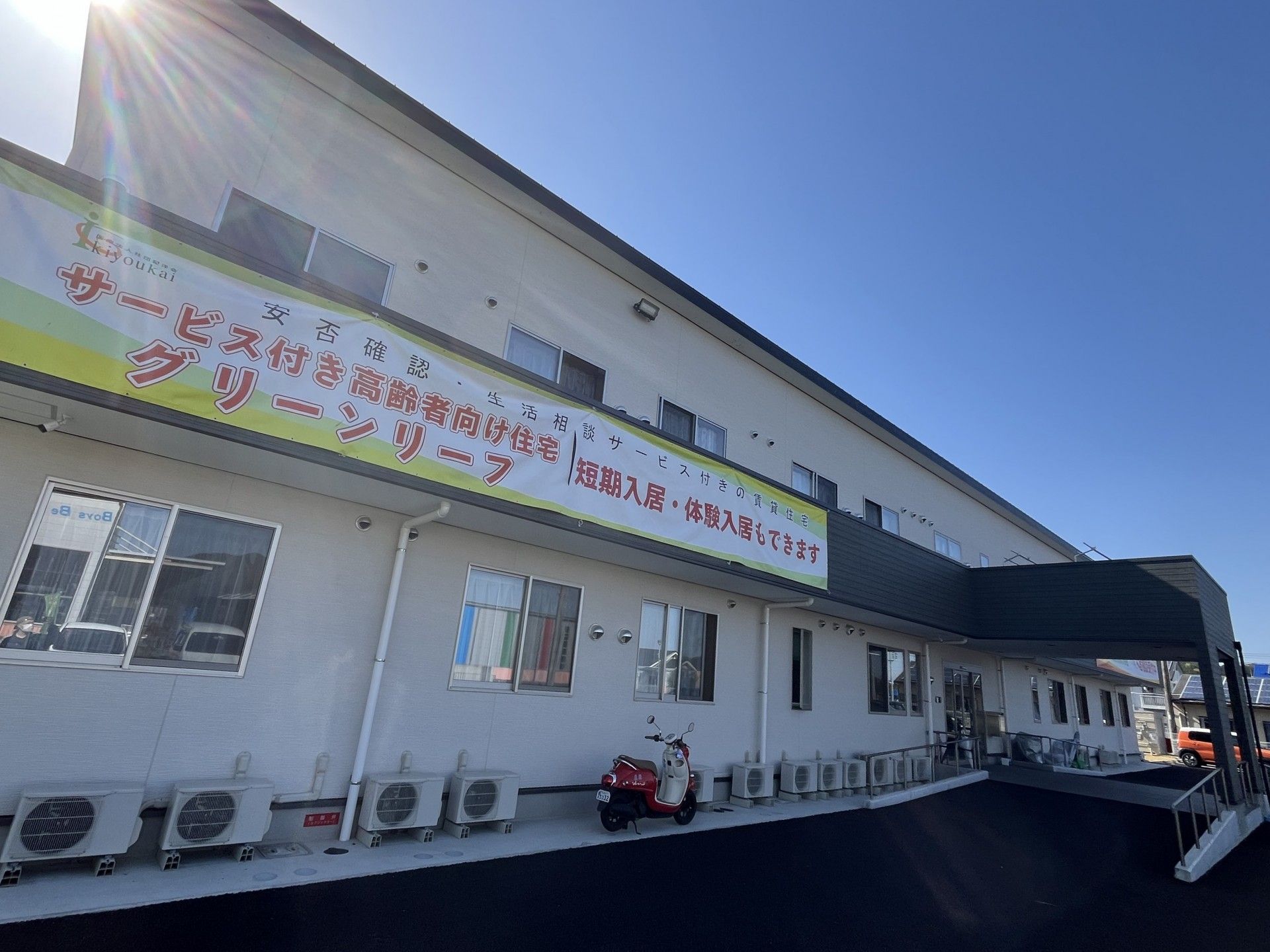遠くを指さしながら笑顔でいる車いすの高齢女性と付き添いのブルーのエプロンを付けた女性スタッフ。