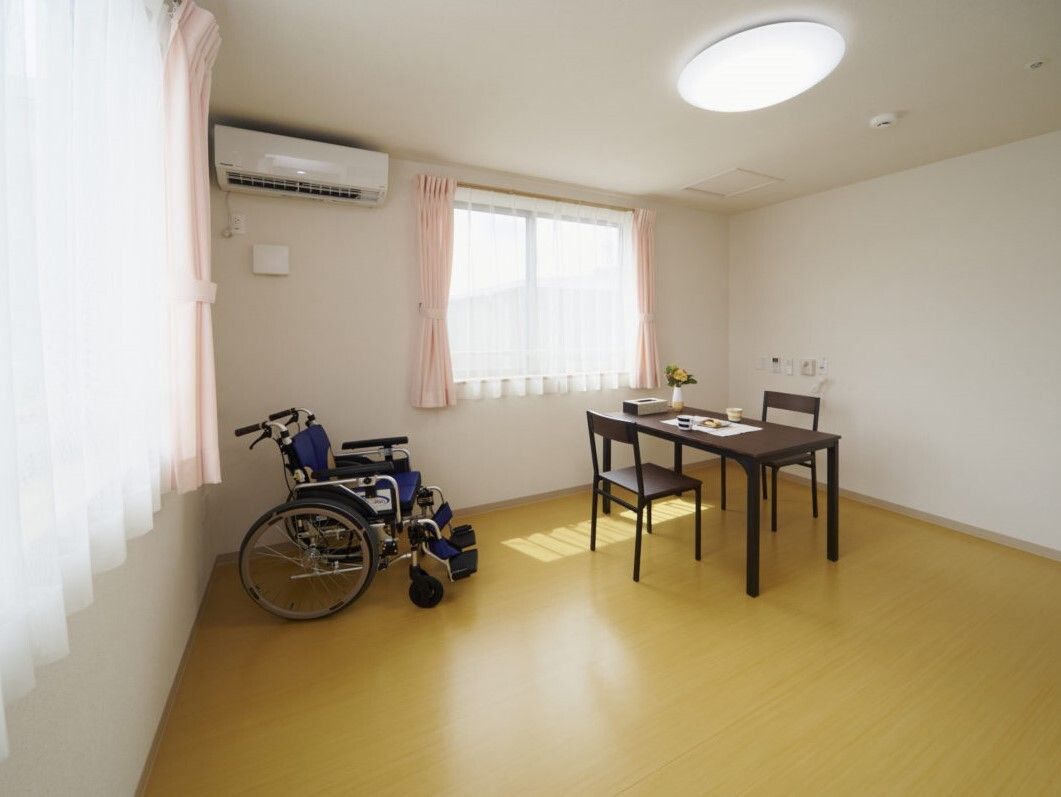 レースカーテンとカーテンが付いた窓がある広い部屋には車いすやテーブルセット、エアコンがある。