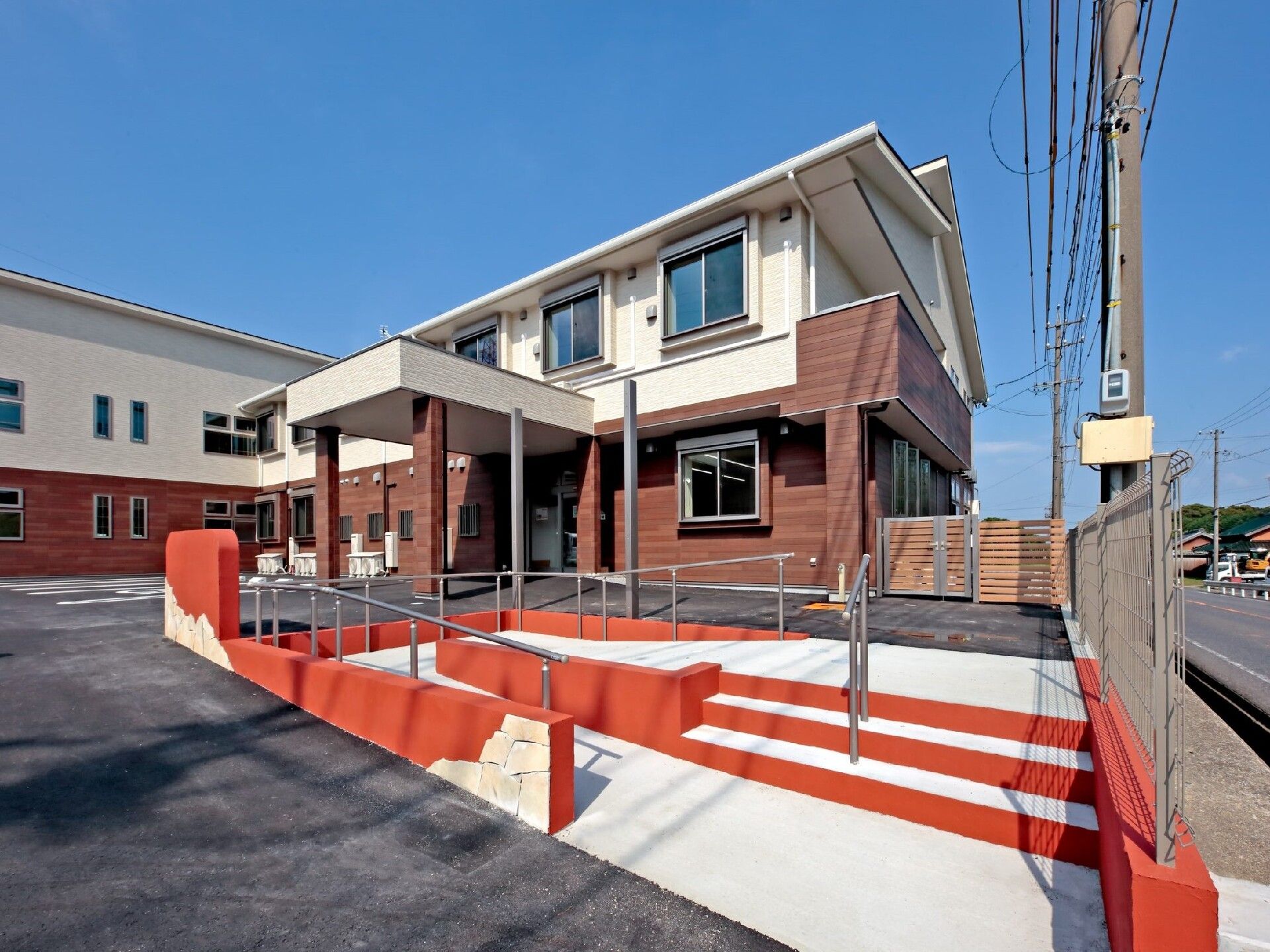 赤と白の手すり付きのスロープと階段が設置されている２階建ての建物は日当たりのいい場所に建っている。