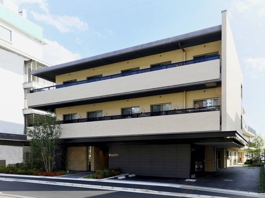 3階建ての建物の各フロアにベランダがあり、建物の周りには緑が植えられ、住人が玄関から入ろうとしている。