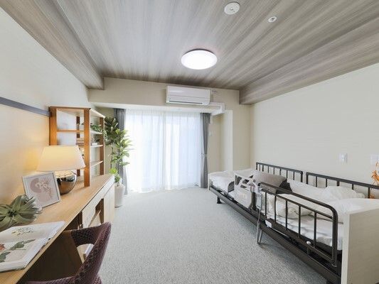 エアコンが付いた居室には寝具付きのベッドや観葉植物、デスクセットがあり、ベッドサイドには緊急通報装置がある。