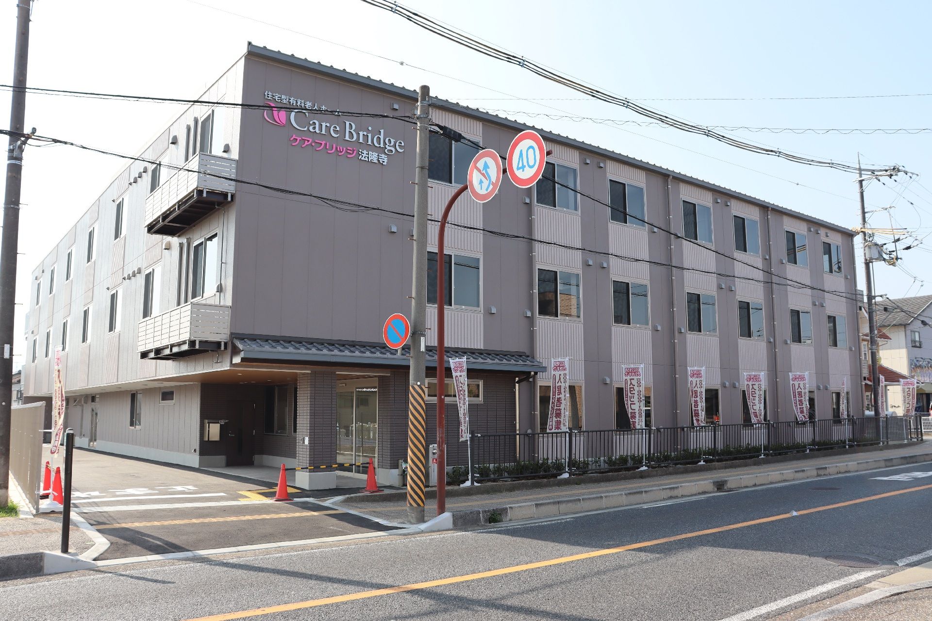 グレー系の外観の3階建ての建物の前に２名の人が歩いていて、建物の前には車が止まっている。外壁にはホーム名が書かれている。