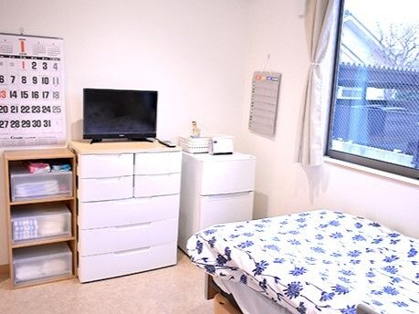 プラスチックの収納タンスやテレビ、冷蔵庫、寝具付きのベッドが置かれた部屋。