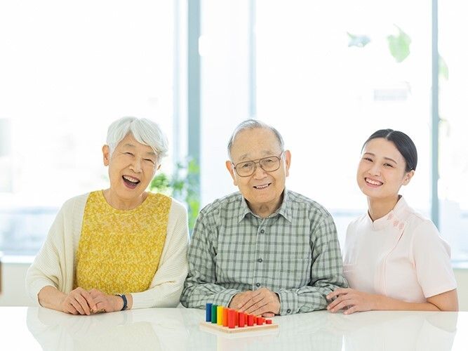 中央に高齢男性、左側に高齢女性、右側に女性スタッフがいて、高齢の男女は座り、スタッフは寄り添い微笑んでいる。