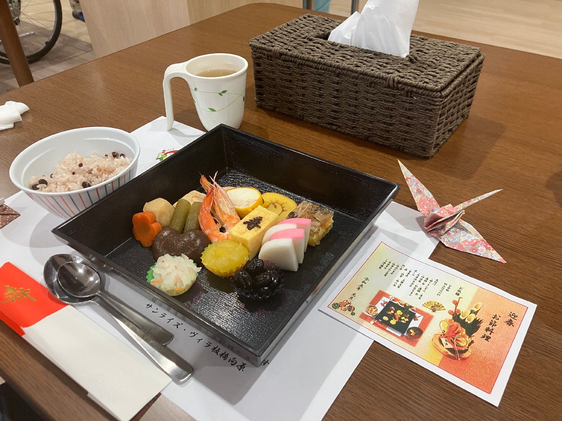 カラフルなお品書きや折り紙の鶴が添えられた黒い重箱の中にはおせち料理が並んでいる。