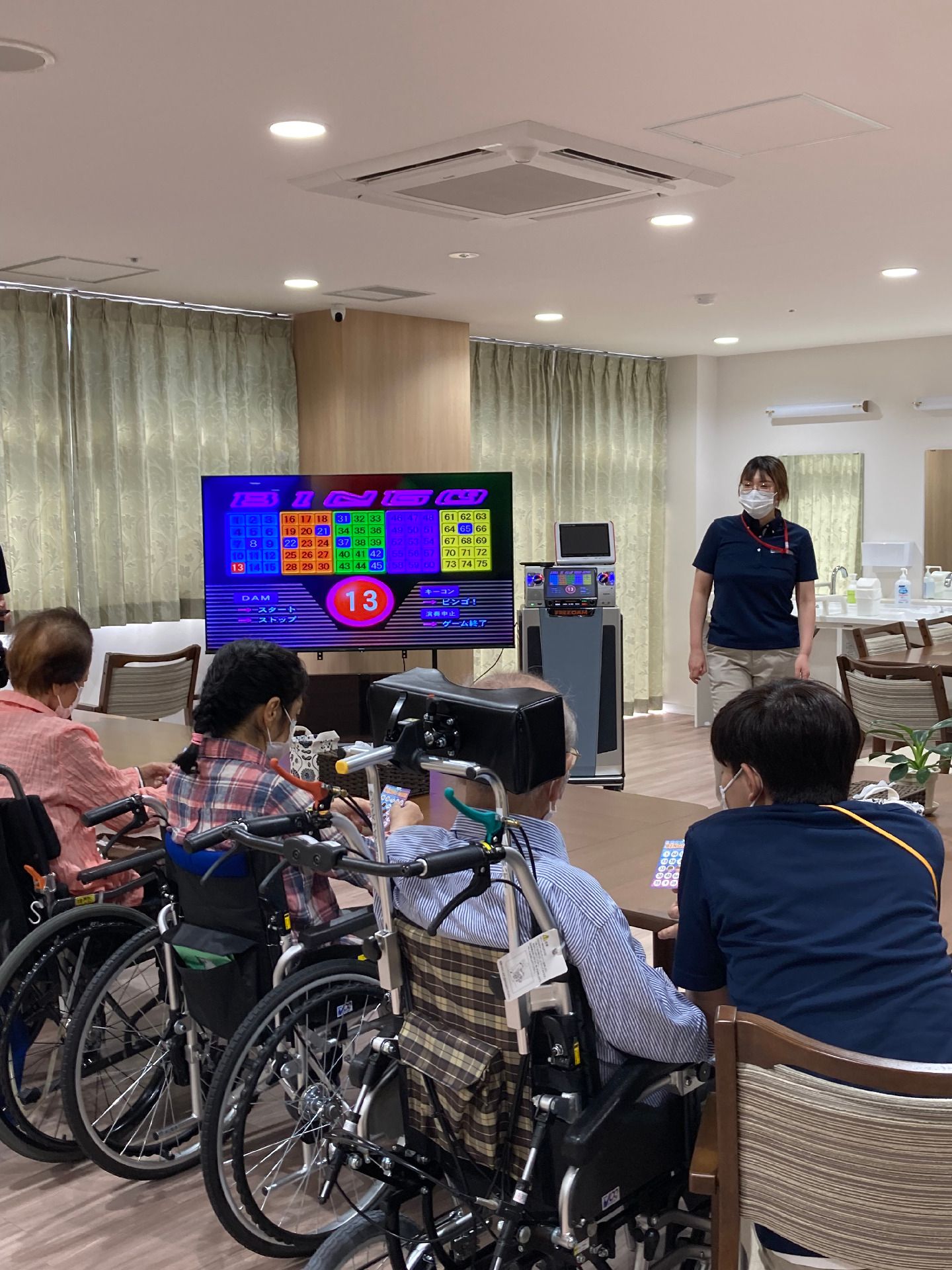 大きなテレビ画面にはゲームのようなものが映っていて、車いすの高齢者たちとスタッフがいる。