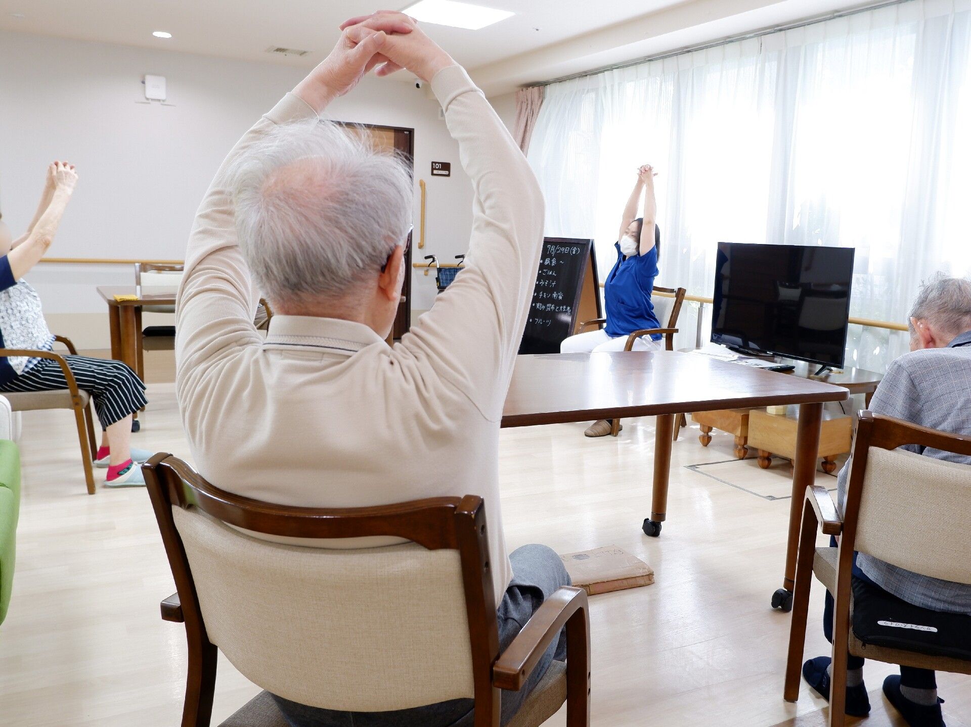 高齢者たちが座ってエクササイズをしている様子。前には女性スタッフがいて手本を見せている。