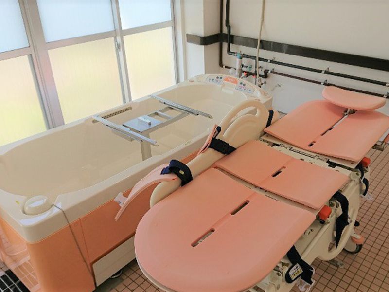 白とピンクの機械浴が置かれた明るい浴室。ストレッチャータイプのリフトと浴槽タイプの機械がある。