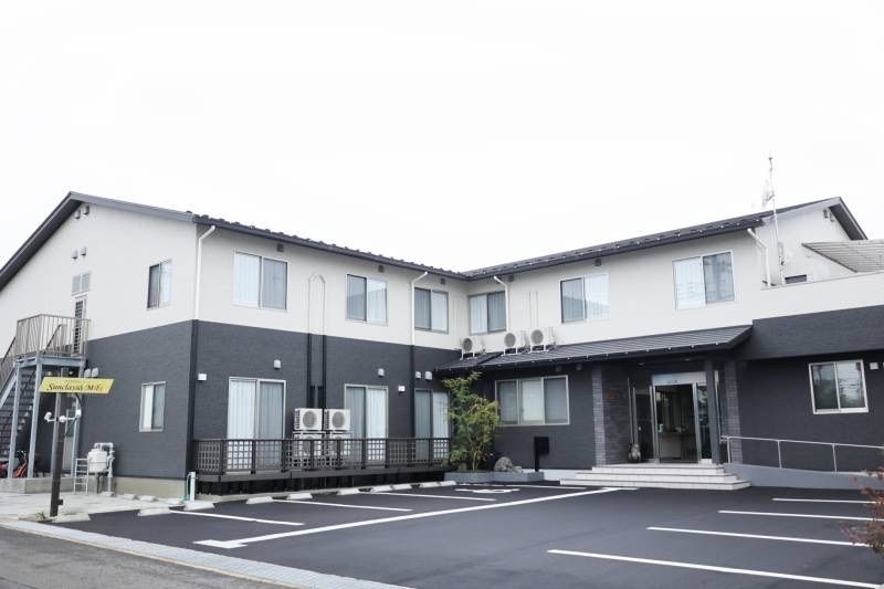 エル字型の２階建ての建物はモノトーンで玄関には屋根があり、スロープと階段も付いている。