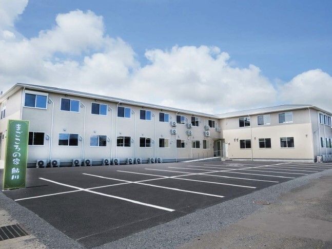 白を基調とした２階建ての建物はエル字型で、敷地内には広い駐車場があり、看板も立っている。