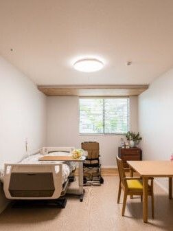 寝具付きのベッドやテーブルセットが置かれたフローリングの部屋。窓のそばには観葉植物が飾られている。