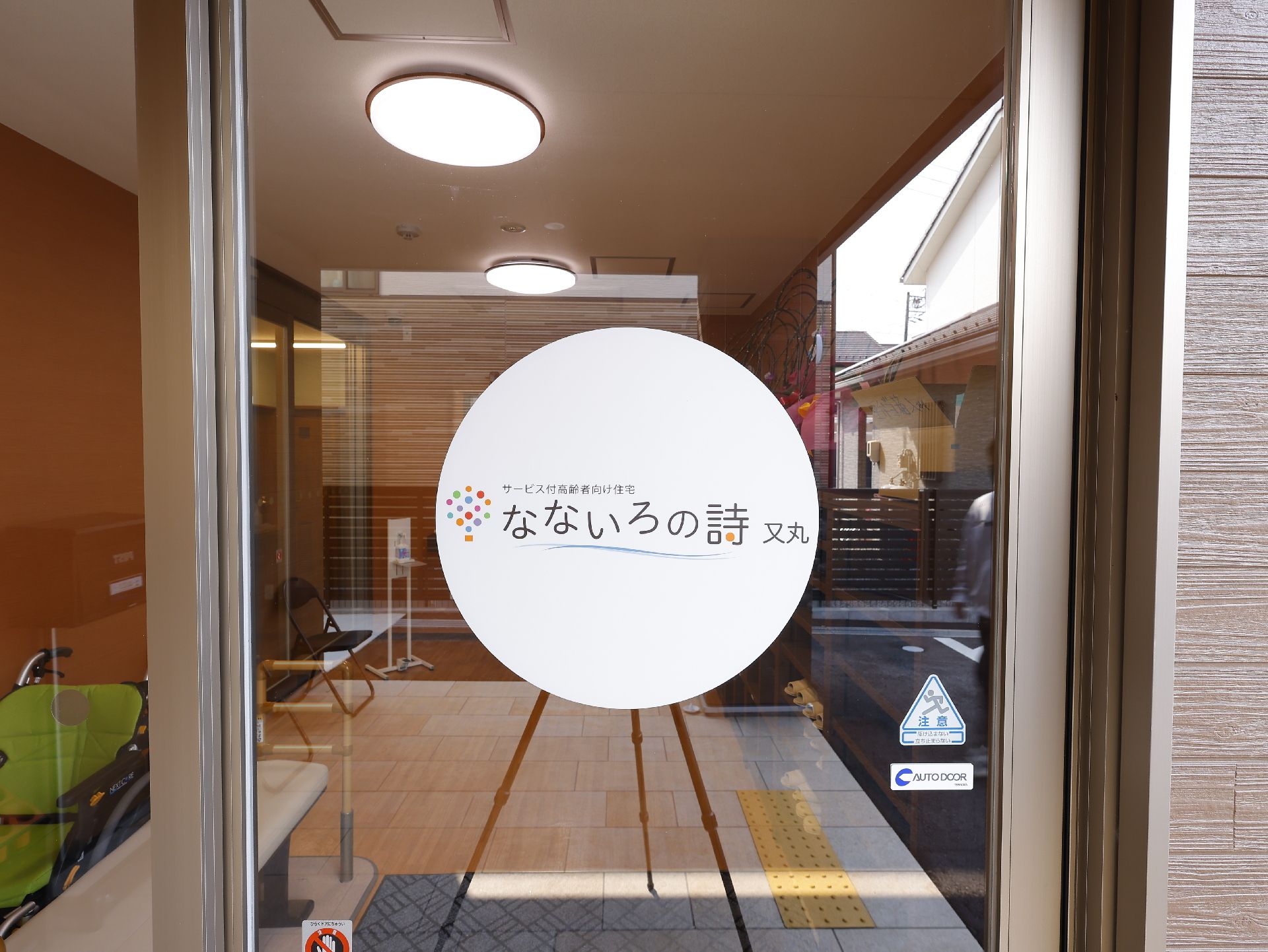 白いゆったりとした服を着た高齢女性が車いすの高齢男性のそばに立ち、笑顔で話している。