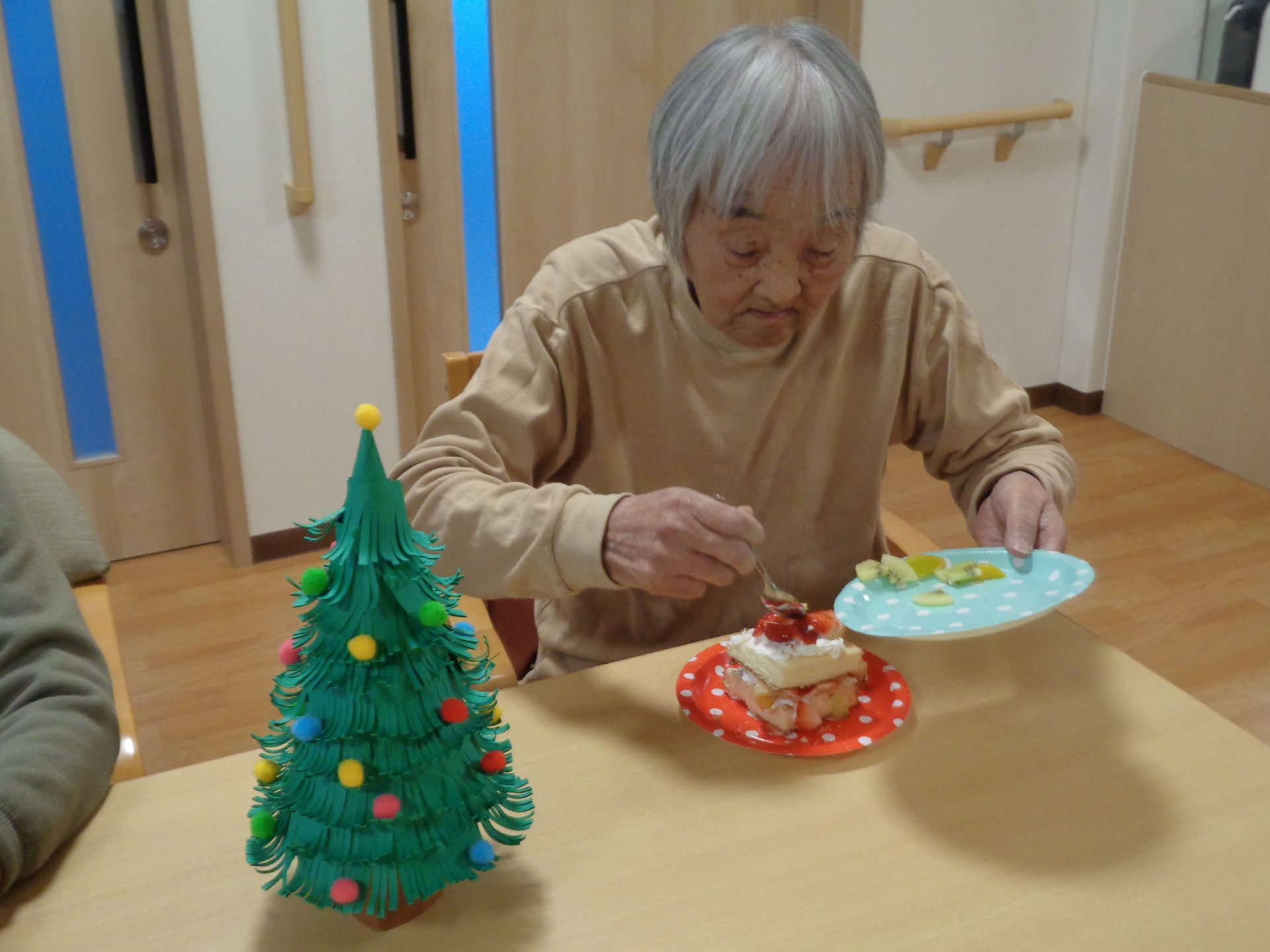 クリスマスツリーが飾られたダイニングテーブルの上にはいちごのショートケーキがあり、高齢女性が食べようとしている。
