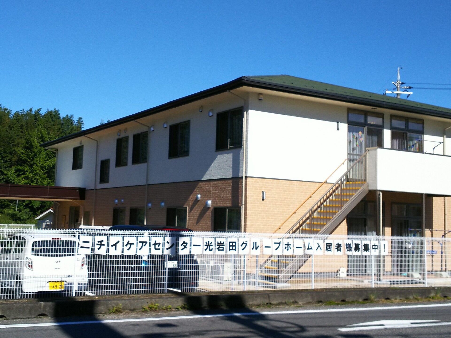 白とベージュの２階建ての建物。屋外階段が設置され、敷地内には駐車場があり、車が止まっている。